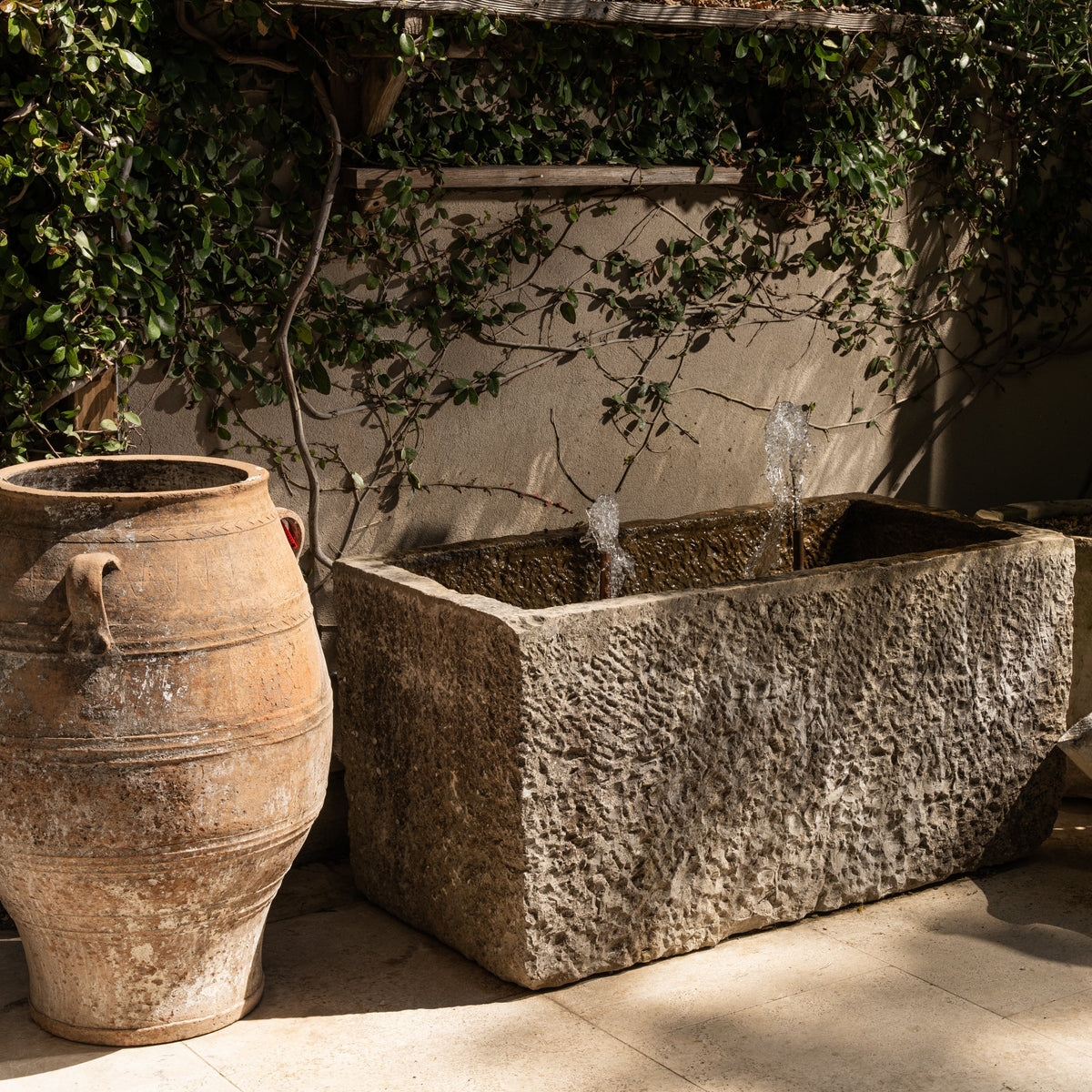 The Well by BDAntiques antique 3-handled terracotta olive jar next to a stone fountain in a garden setting.