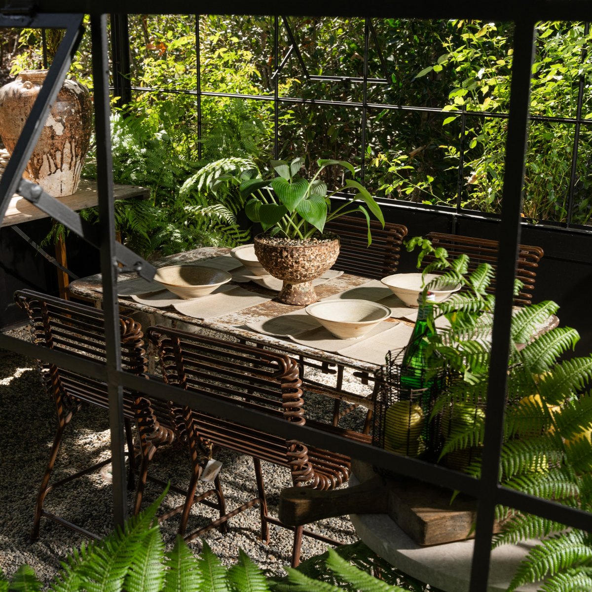 The Well by BDAntiques rust-colored iron rod outdoor chairs arranged around a dining table in a sunlit greenhouse.