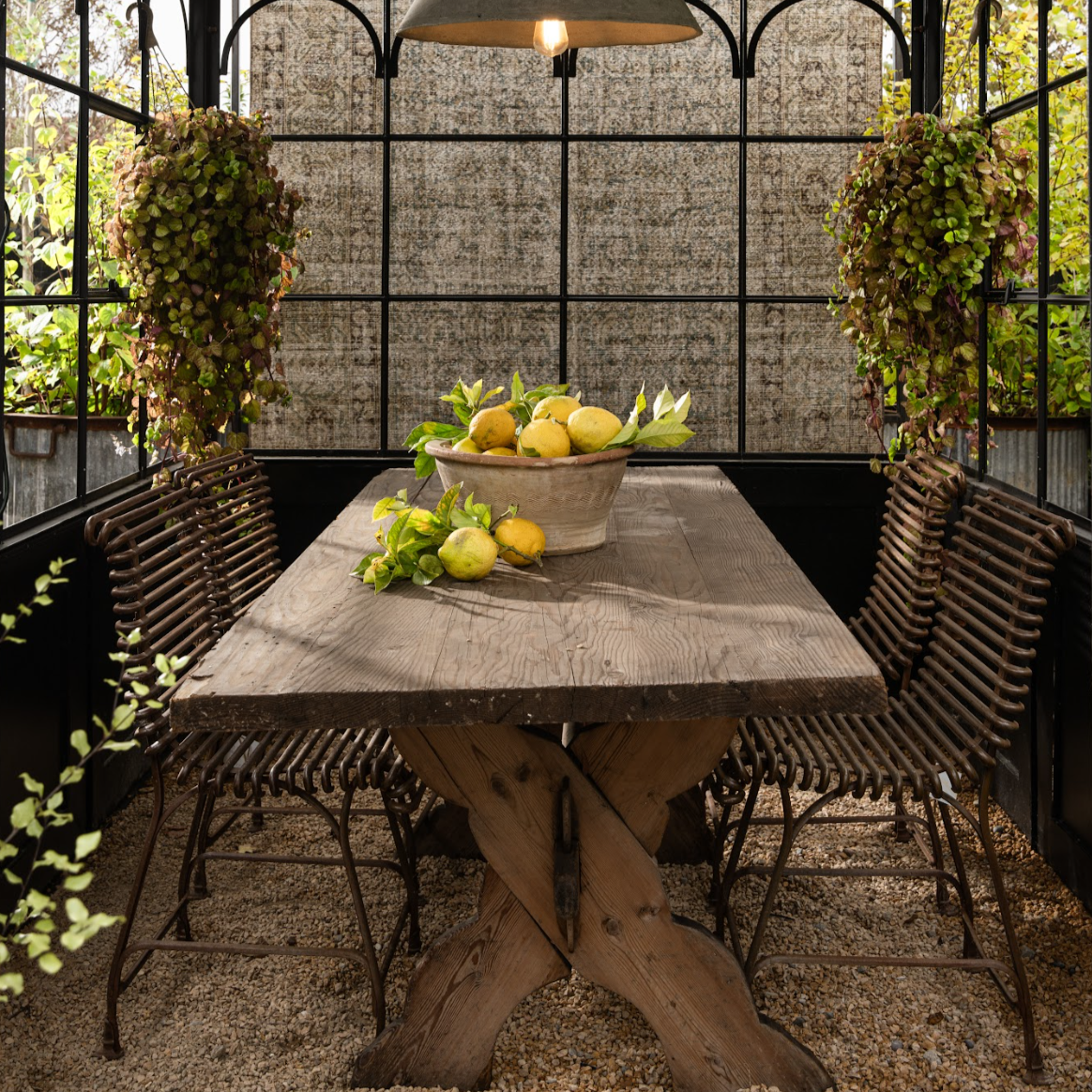 The Well by BDAntiques rust-colored metal outdoor chairs with curved iron rod design in a greenhouse dining setting.